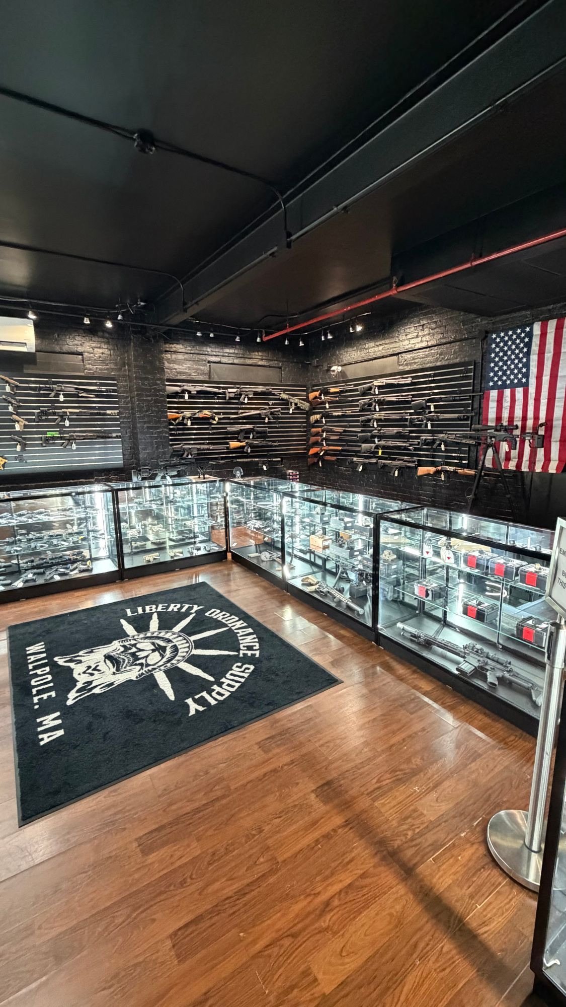The Liberty Ordnance Supply sales floor in Walpole: lit glass cases, slatwalls of rifles and shotguns on black brick, an American flag, and the store's logo rug